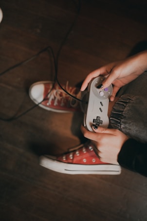 Hands holding a retro game controller, with visible red sneakers and a wooden floor in the background.