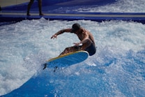 A person is performing a trick on a surfboard in a wave pool, showcasing agility and balance. Water splashes around as the board is skillfully maneuvered mid-air. The bright blue hues of the water contrast with the figure's tanned skin and dark swimwear.