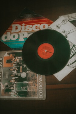 A vinyl record with a red label sits prominently on top of several album covers, which include colorful graphics and illustrations. The covers exhibit a retro design style, typical of older record albums, with one displaying a band in performance.