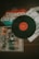 A vinyl record with a red label sits prominently on top of several album covers, which include colorful graphics and illustrations. The covers exhibit a retro design style, typical of older record albums, with one displaying a band in performance.