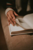 An elegant close-up of a hand turning the pages of a textured, off-white book with gold accents.