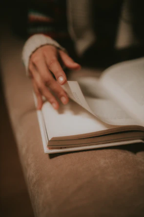 An elegant close-up of a hand turning the pages of a textured, off-white book with gold accents.