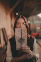 A person with long hair and glasses is holding a map with an intricate, old-fashioned design. The map has a castle illustration and is somewhat worn, suggesting a vintage style. The person is standing in a warmly lit setting with wooden walls and a blurred background.