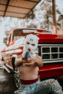 A person wearing a white ski mask with dollar signs on it is holding a blue item pointed forward. They are kneeling in front of a red vintage truck. The person is wearing a sleeveless top and ripped blue jeans. The background shows an open structure with trees visible through it.