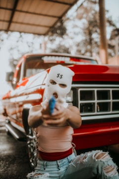 A person wearing a white ski mask with dollar signs on it is holding a blue item pointed forward. They are kneeling in front of a red vintage truck. The person is wearing a sleeveless top and ripped blue jeans. The background shows an open structure with trees visible through it.