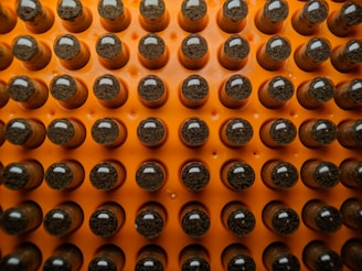 A grid of reflective capsules or vials with dark material inside, arranged on an orange background. They are organized in neat rows, with each having a shiny, rounded top.