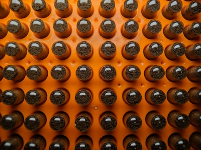A grid of reflective capsules or vials with dark material inside, arranged on an orange background. They are organized in neat rows, with each having a shiny, rounded top.