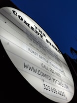 A white, oval-shaped marquee sign displays information about comedy events and contact details. The sign is illuminated against a deep blue, possibly evening sky.