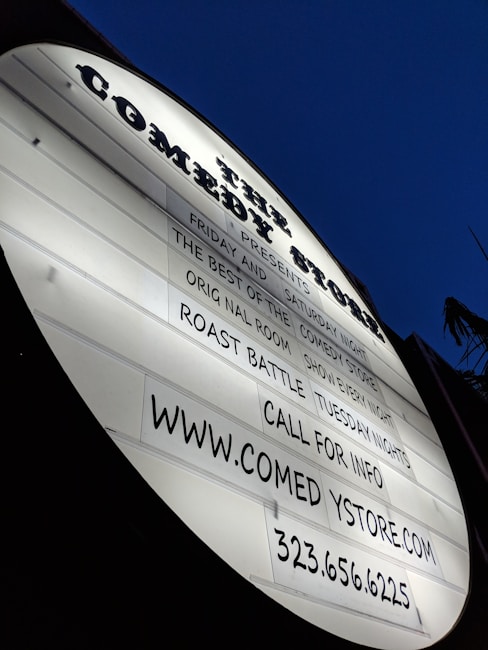 A white, oval-shaped marquee sign displays information about comedy events and contact details. The sign is illuminated against a deep blue, possibly evening sky.