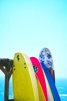 Brightly colored surfboards are propped up against a wooden structure with a clear blue sky and ocean in the background. The surfboards are in shades of yellow, white, pink, and blue, with various designs and logos visible on them.