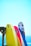 Brightly colored surfboards are propped up against a wooden structure with a clear blue sky and ocean in the background. The surfboards are in shades of yellow, white, pink, and blue, with various designs and logos visible on them.
