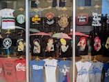 A storefront display of T-shirts, featuring various designs. The shirts depict famous characters with a twist, Mexican cultural symbols, and humorous or bold statements. There's a blend of black, white, and colorful tees, some with logos and others with illustrations. The reflection of the street and people can be seen in the glass.
