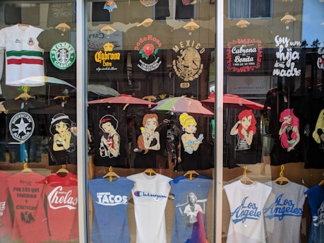 A storefront display of T-shirts, featuring various designs. The shirts depict famous characters with a twist, Mexican cultural symbols, and humorous or bold statements. There's a blend of black, white, and colorful tees, some with logos and others with illustrations. The reflection of the street and people can be seen in the glass.