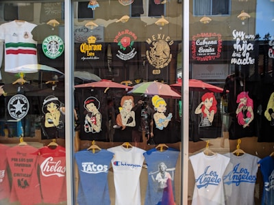 A storefront display of T-shirts, featuring various designs. The shirts depict famous characters with a twist, Mexican cultural symbols, and humorous or bold statements. There's a blend of black, white, and colorful tees, some with logos and others with illustrations. The reflection of the street and people can be seen in the glass.