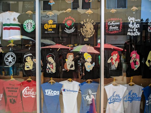 A storefront display of T-shirts, featuring various designs. The shirts depict famous characters with a twist, Mexican cultural symbols, and humorous or bold statements. There's a blend of black, white, and colorful tees, some with logos and others with illustrations. The reflection of the street and people can be seen in the glass.