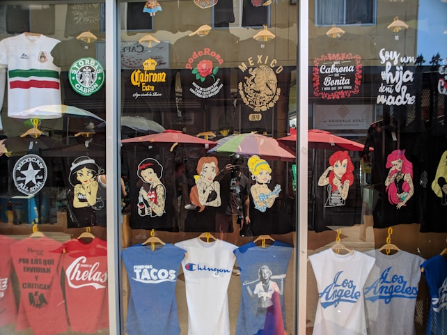 A storefront display of T-shirts, featuring various designs. The shirts depict famous characters with a twist, Mexican cultural symbols, and humorous or bold statements. There's a blend of black, white, and colorful tees, some with logos and others with illustrations. The reflection of the street and people can be seen in the glass.