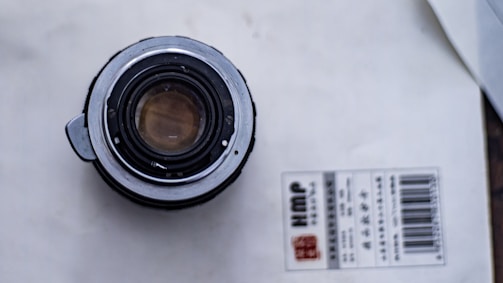 A camera lens is placed on a white surface next to a packaging label with text and a barcode. The lens is viewed from the back, showing the metal and glass elements.