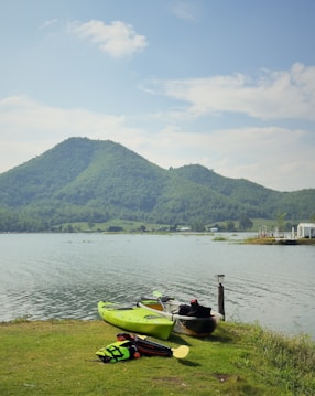 A tranquil lakeside scene features two kayaks on a grassy shore, with one kayak in bright green and another in a more muted color. Nearby, a life jacket and paddle are placed on the grass. The lake is calm, nestled in a valley surrounded by lush, green mountains beneath a clear, blue sky with scattered clouds. In the distance, buildings are faintly visible along the shoreline.