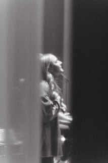A black and white image captures a person with long hair, possibly a woman, in a side profile. The individual appears to be singing or speaking passionately. The image has a soft focus, creating a dreamy and ethereal atmosphere.