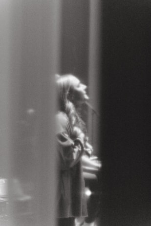 A black and white image captures a person with long hair, possibly a woman, in a side profile. The individual appears to be singing or speaking passionately. The image has a soft focus, creating a dreamy and ethereal atmosphere.