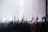 A large crowd of people with raised hands is watching a musical performance. The stage features silhouettes of band members with instruments, under bright stage lights, creating an energetic atmosphere.