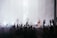 A large crowd of people with raised hands is watching a musical performance. The stage features silhouettes of band members with instruments, under bright stage lights, creating an energetic atmosphere.