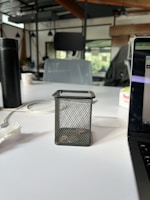 A close-up view of a workspace, featuring a mesh pen holder with coins inside placed on a white table. On the left, there's a black insulated bottle and a crumpled tissue. On the right, part of an open laptop with a visible screen edge is seen. The background reveals an open-plan office environment with chairs, desks, and hanging lights, creating a casual, contemporary office setting.