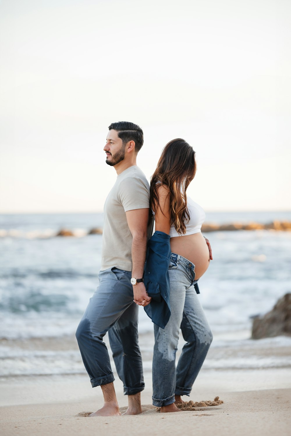 a pregnant couple standing on the beach with the ocean in the background