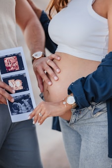 A person wearing a white top and jeans is holding their belly, indicating pregnancy. Another person, partially shown, holds ultrasound images. Both display watches, and one also wears a bracelet.