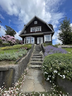 A large house with a dark exterior featuring multiple windows and a steeply pitched roof. A stone pathway leads up to the entrance, bordered by neatly arranged greenery and colorful flowering plants. The sky above is partly cloudy, offering a contrast to the vivid green vegetation and the house's dark facade.