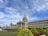 A grand government building with intricate architectural details, featuring large columns and a dome. The structure is surrounded by neatly maintained gardens with various shrubs and trees. A clear blue sky with soft, scattered clouds forms the backdrop.