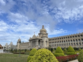 A grand government building with intricate architectural details, featuring large columns and a dome. The structure is surrounded by neatly maintained gardens with various shrubs and trees. A clear blue sky with soft, scattered clouds forms the backdrop.