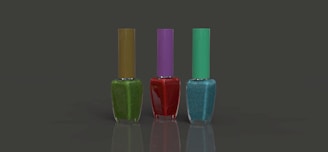 Three nail polish bottles are arranged in a row against a plain dark background. The bottles have different colored polish: green with an olive cap, red with a purple cap, and blue with a teal cap. Each bottle is rectangular with a slender cap, and the surface is reflective.