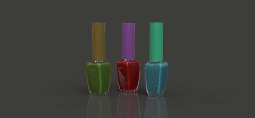 Three nail polish bottles are arranged in a row against a plain dark background. The bottles have different colored polish: green with an olive cap, red with a purple cap, and blue with a teal cap. Each bottle is rectangular with a slender cap, and the surface is reflective.