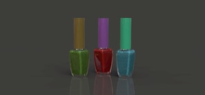 Three nail polish bottles are arranged in a row against a plain dark background. The bottles have different colored polish: green with an olive cap, red with a purple cap, and blue with a teal cap. Each bottle is rectangular with a slender cap, and the surface is reflective.