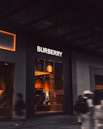 An upscale storefront with a prominent 'Burberry' sign above large display windows. Inside, warm lighting highlights fashionable clothing displays and mannequins. The exterior is dark, contrasting with the bright interior. A few blurred figures, possibly pedestrians, are visible in the foreground, suggesting motion.