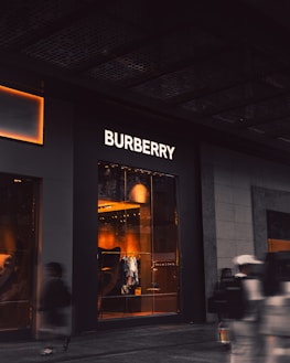 An upscale storefront with a prominent 'Burberry' sign above large display windows. Inside, warm lighting highlights fashionable clothing displays and mannequins. The exterior is dark, contrasting with the bright interior. A few blurred figures, possibly pedestrians, are visible in the foreground, suggesting motion.