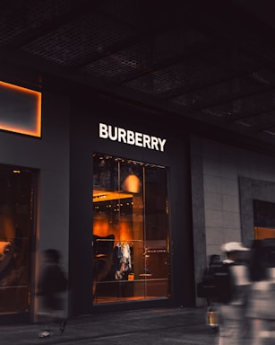 An upscale storefront with a prominent 'Burberry' sign above large display windows. Inside, warm lighting highlights fashionable clothing displays and mannequins. The exterior is dark, contrasting with the bright interior. A few blurred figures, possibly pedestrians, are visible in the foreground, suggesting motion.