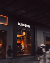 An upscale storefront with a prominent 'Burberry' sign above large display windows. Inside, warm lighting highlights fashionable clothing displays and mannequins. The exterior is dark, contrasting with the bright interior. A few blurred figures, possibly pedestrians, are visible in the foreground, suggesting motion.
