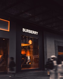 An upscale storefront with a prominent 'Burberry' sign above large display windows. Inside, warm lighting highlights fashionable clothing displays and mannequins. The exterior is dark, contrasting with the bright interior. A few blurred figures, possibly pedestrians, are visible in the foreground, suggesting motion.
