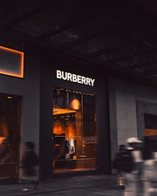 An upscale storefront with a prominent 'Burberry' sign above large display windows. Inside, warm lighting highlights fashionable clothing displays and mannequins. The exterior is dark, contrasting with the bright interior. A few blurred figures, possibly pedestrians, are visible in the foreground, suggesting motion.