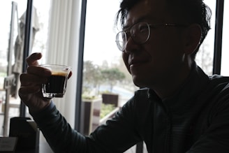 A smiling person holding a coffee cup, sitting in front of a bright window with plants.