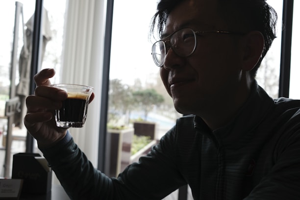 A smiling person holding a coffee cup, sitting in front of a bright window with plants.
