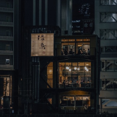 A cozy, urban restaurant is illuminated at night, revealing multiple floors through large windows. The warm, inviting glow from the lights inside contrasts with the surrounding dark city buildings. A sign with Japanese characters is prominently displayed on the building's exterior.