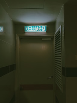 An illuminated exit sign displays the word 'KELUAR' in bold letters with an accompanying running figure symbol. The sign is mounted above a closed door, suggesting an emergency exit. The surrounding area is dimly lit, with a muted green color scheme dominating the scene. The door features a simple silver handle, and there is a louvered panel nearby.