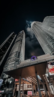 Tall, illuminated skyscrapers stretch upwards against a dark night sky. A prominent building sign reads 'Suria KLCC', and lights from various windows create a striking urban scene. Clouds are faintly visible, adding depth to the moody atmosphere.