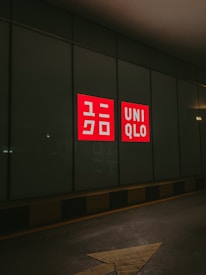 A dark street with a large glass window featuring a bright red and white UNIQLO logo, illuminated against the dark surroundings. The arrow painted on the pavement in the foreground points towards the glass wall.