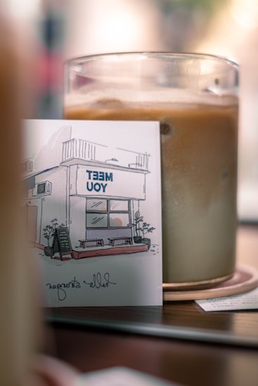 A glass cup filled with an iced beverage sits on a wooden table. Next to the cup, there is a small card featuring an illustration of a storefront with the words 'MEET YOU'. The card includes a sketch of a building with plants and a small chalkboard sign out front.