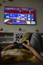 A person is lounging on a couch, holding a Sony game controller, playing a video game displayed on a wall-mounted TV screen. The game has bright and colorful graphics with red and blue tones. The room includes decorative elements like shelves, small potted plants, and wall-mounted ornaments.