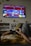 A person is lounging on a couch, holding a Sony game controller, playing a video game displayed on a wall-mounted TV screen. The game has bright and colorful graphics with red and blue tones. The room includes decorative elements like shelves, small potted plants, and wall-mounted ornaments.